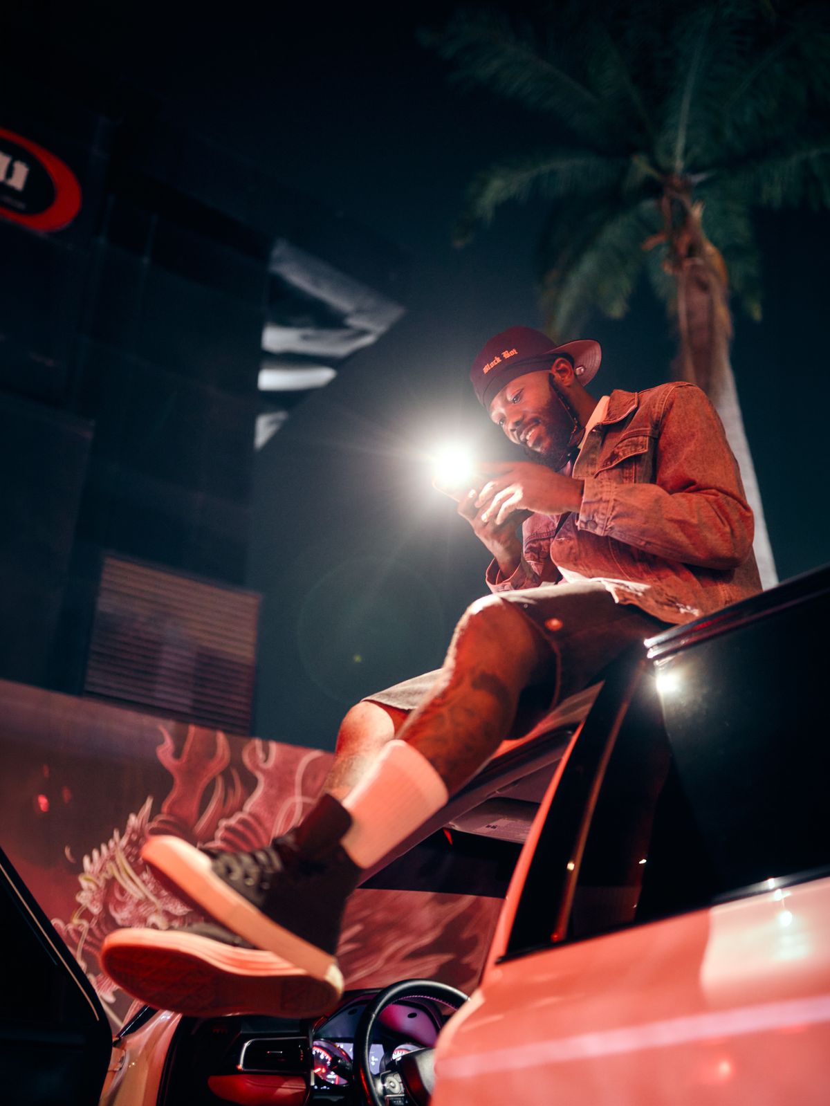 DJ Romie smiling, lit by phone light at night beside a car