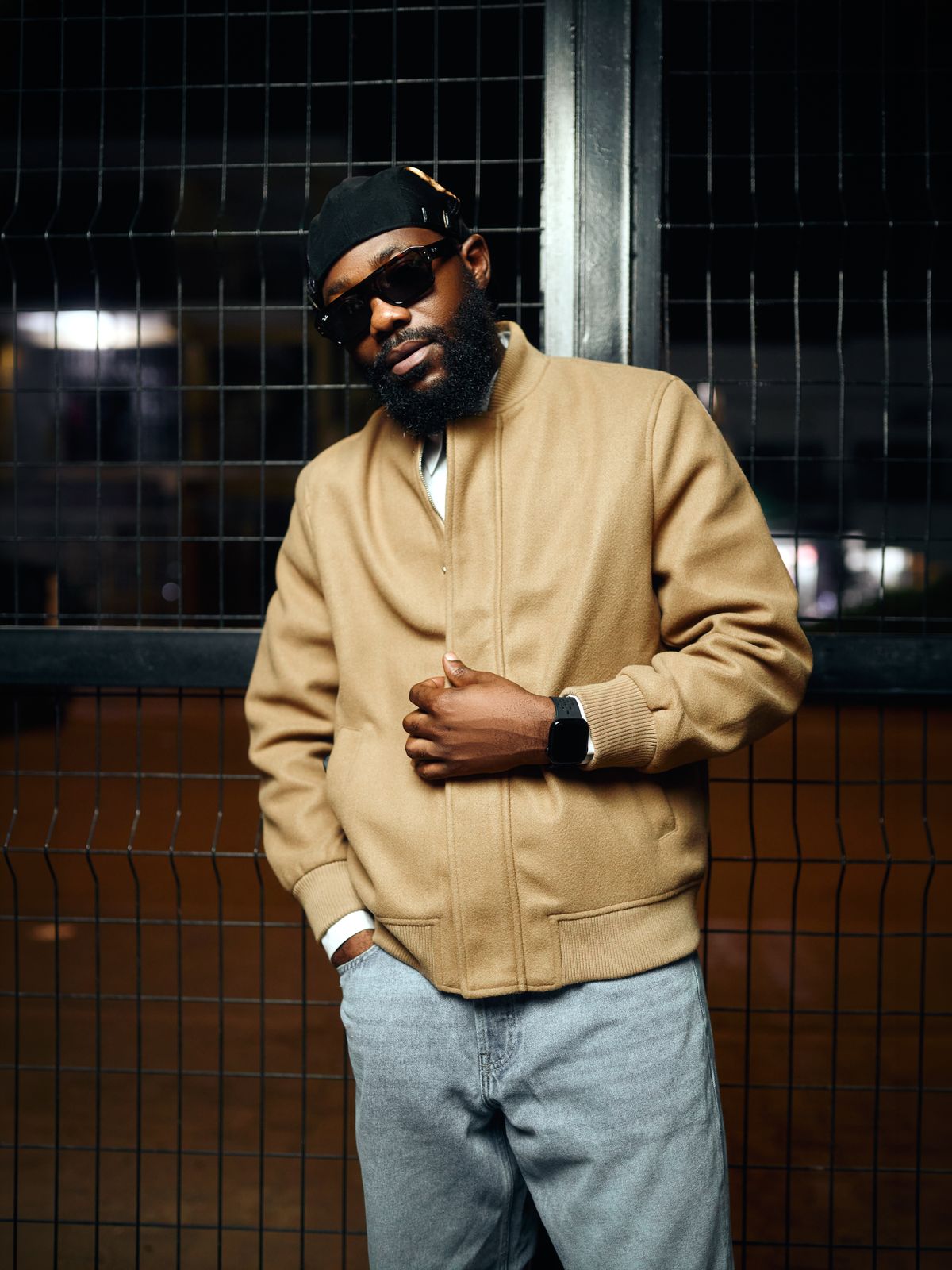 DJ Romie in a tan bomber jacket against a metal fence at night